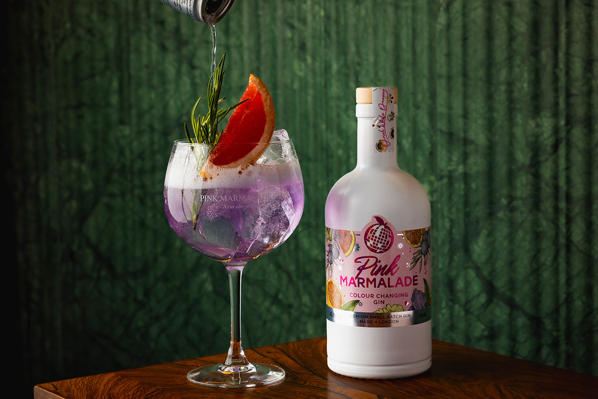 Pink Marmalade Gin on a table next to a bright pink purple Gin & Sonic cocktail with grapefruit garnish and tonic pouring