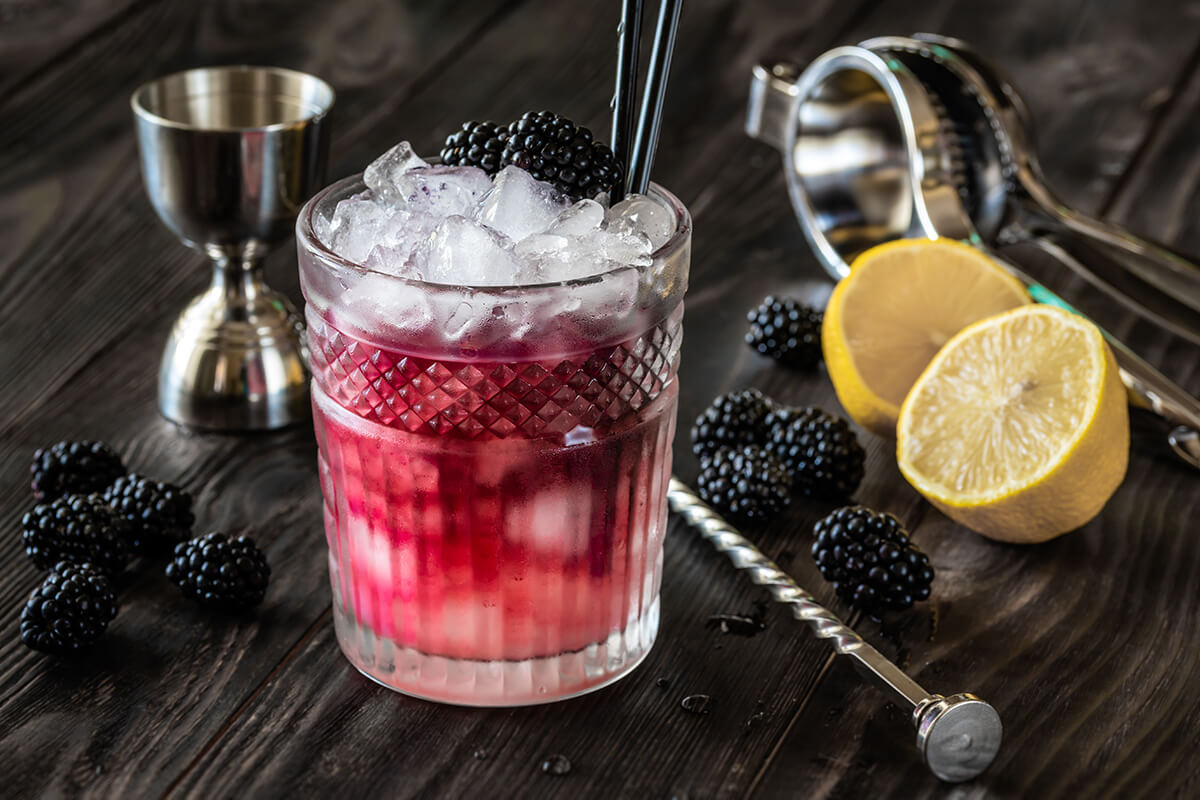 Gin Bramble cocktail in a glass with ice and blackberry garnish, on a table with cocktail stirrer and fresh lemon